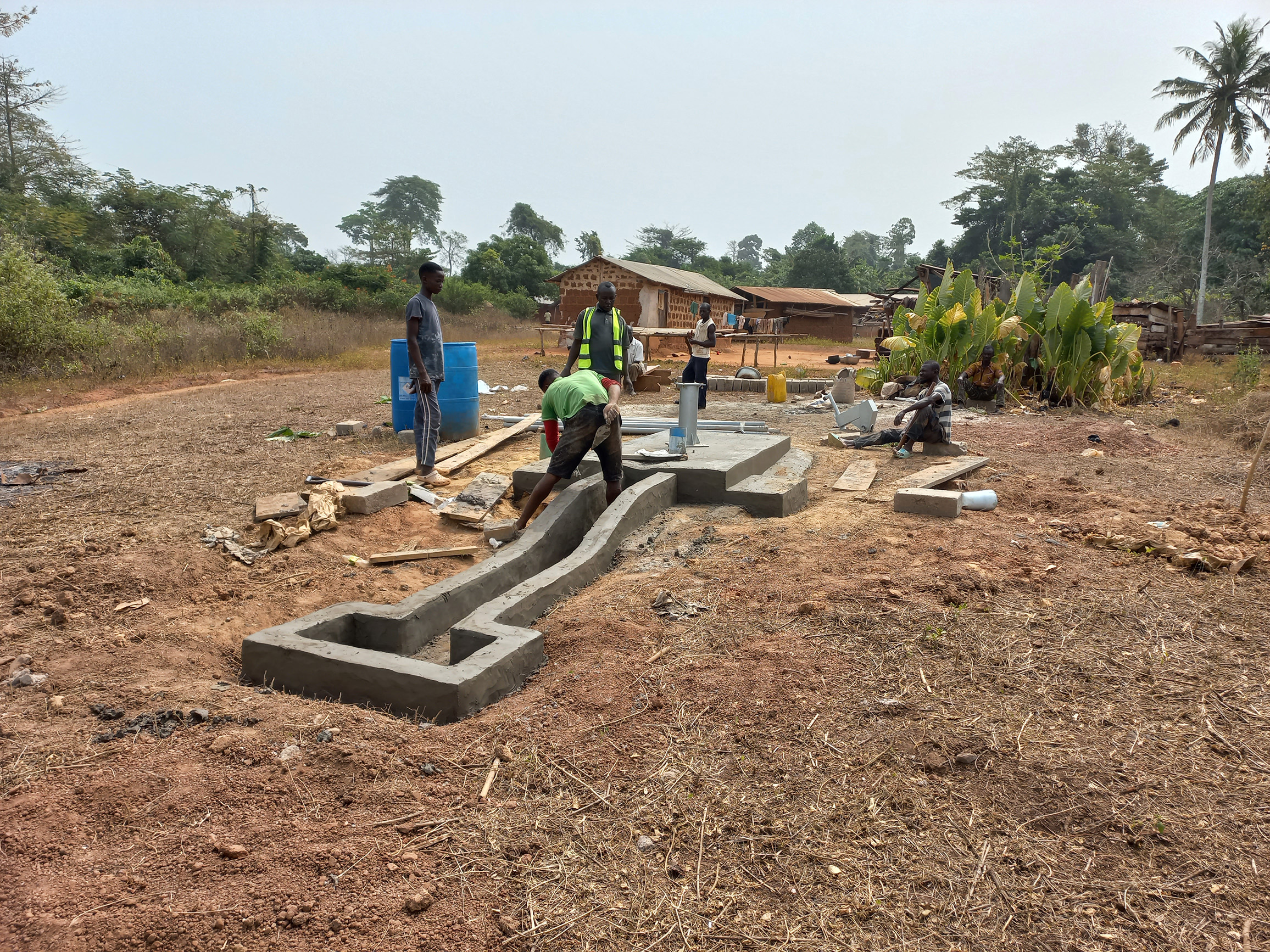 Mehrere Bauarbeiter arbeiten an einer neuen Brunnenanlage in einer ländlichen Region Ghanas. Im Vordergrund verstreicht ein Arbeiter frischen Beton an einer langen Abflussrinne, die zu einem quadratischen Auffangbecken führt.