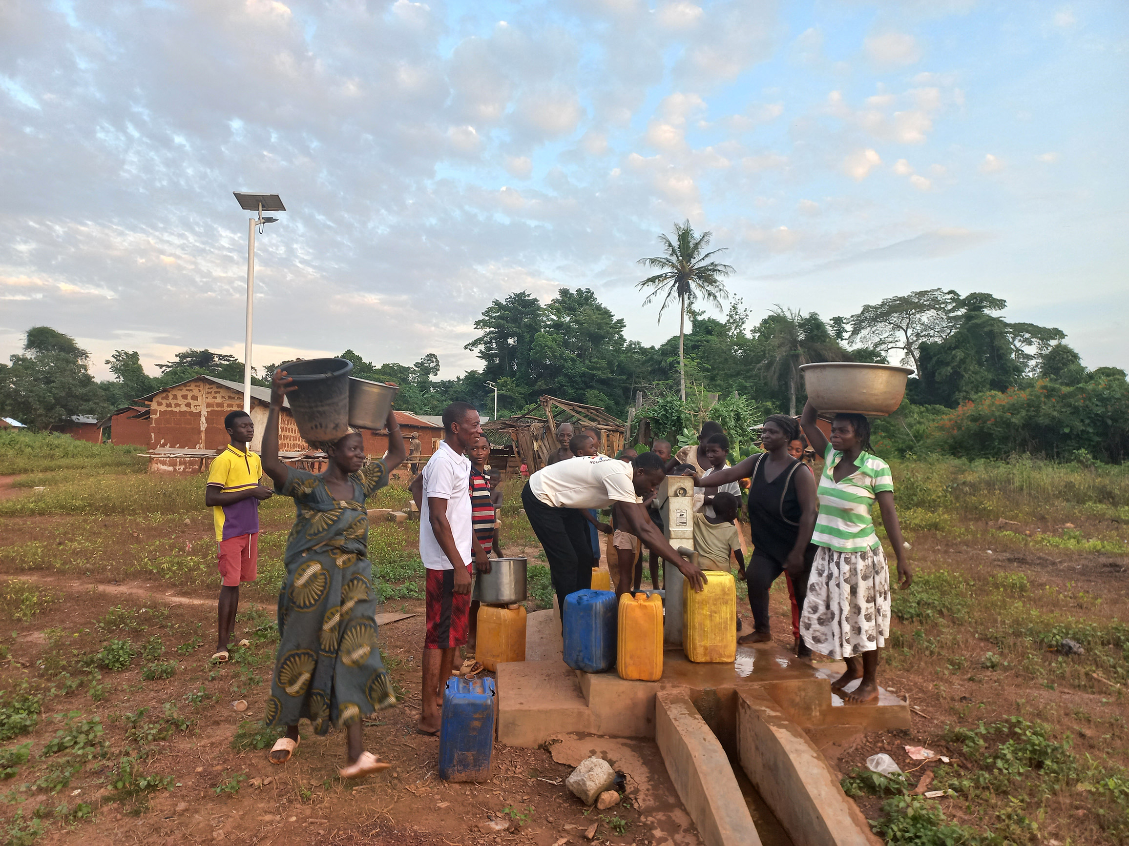 Eine belebte Szene an der fertiggestellten Brunnenanlage in Ghana. Mehrere Frauen und Männer füllen Eimer und gelbe Kanister an der Handpumpe auf. Im Hintergrund ist die installierte Beleuchtungsanlage zu sehen.
